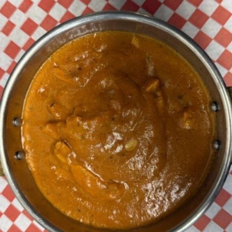 A metal bowl filled with rich, orange Indian curry, served on a red and white checkered paper.