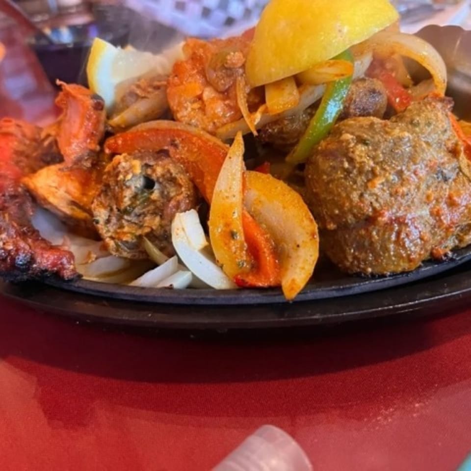 Sizzling Indian platter with chicken, shrimp, onions, peppers, and lemon wedges on a black plate.